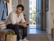 Man pauses with his phone beside a bag of groceries in an apartment entrance.