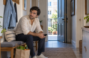 Man pauses with his phone beside a bag of groceries in an apartment entrance.